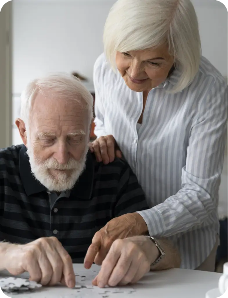Un homme âgé avec une barbe blanche et une femme aux cheveux blancs travaillent ensemble sur un puzzle blanc. La femme pose une main encourageante sur l'épaule de l'homme, illustrant le soutien aux seniors confrontés à la maladie d'Alzheimer avec l'association Le Connétable à Dinan.